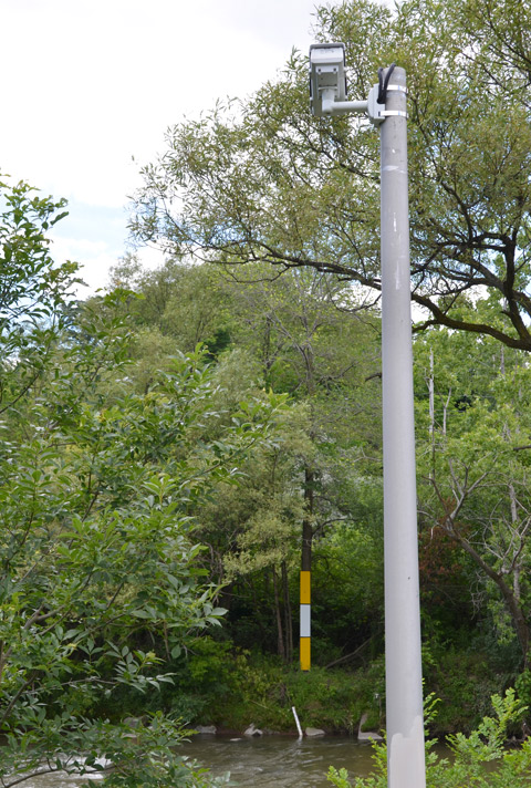 surveillance camera on a tall pole, aimed at rulers and markers on the far side of a river, keeping an eye on the water level 