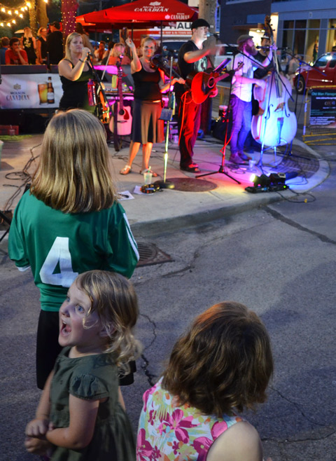 three young girls watch an evening outdoor music performance by a band of 6 musicians on a street corner