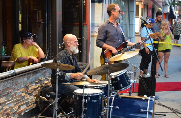 a woman is seated in a restaurant by the window that is open, she is looking out and watching a group of musicians perform, there is a man on a drum set as well as another man. 