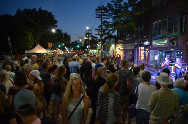 evening, darkening sky, many people on the street at a street festival, most people are facing away from the camera except one woman in front who is looking straight ahead. Musicians playing on the right, standing in front of stores 