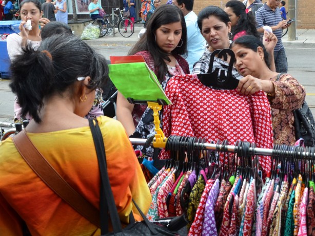 three women are looking at a blouse (dress?), red and white pattern, on a hanger at an outdoor sidewalk sale, as part of a street festival. They are looking at the same piece of clothing