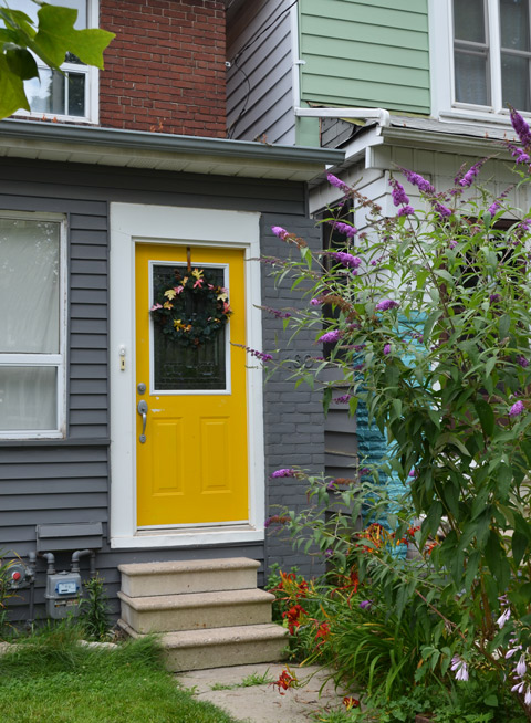 yellow front door on a greyhouse, with lots of flowers in front including purple butterfly bush 