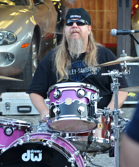 bearded man with sunglasses and black basebell cap sits at a drum set. Hhis baseball cap says Zedhead on it. 
