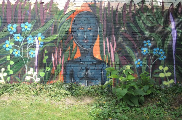 mural on a wall with green plants growing in front, mural is a blue person from the chest up, no hair. Some plants are part of the mural too.