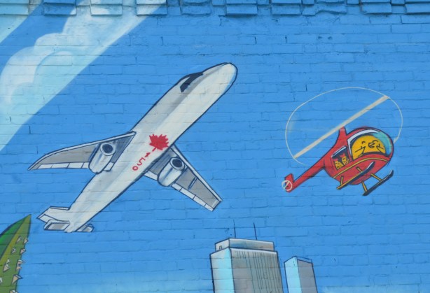 part of a large mural, blue sky painted background, the underside of an airplane as it passes overhead, painted with 150 and a red maple leaf, flying beside it is a red helicopter with a Uber5000 birdie flying it. 