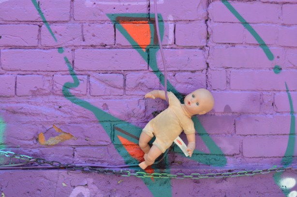 purple wall, graffiti onthe wall, also a chain runs horizontally across the bottom of the picture. A toy doll with no clothes has its arm tucked into a green wire. 