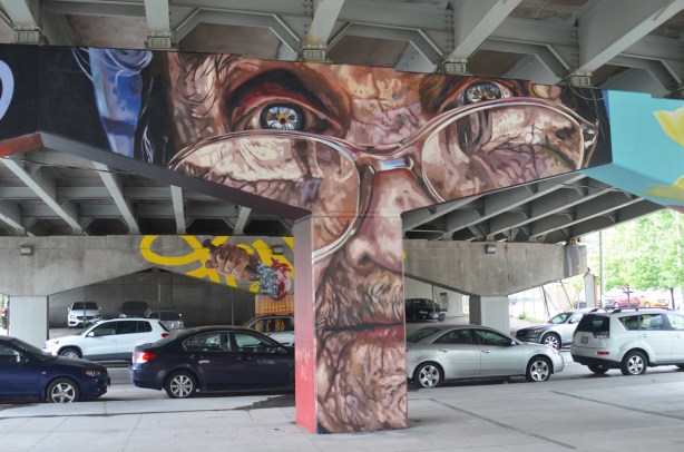 cars parked under a bent that has been painted with a wrinkly brown man's face wearing glasses