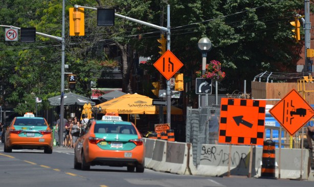 street scene, two orange and green taxis drive by a construction site with orange traffic signs, arrows saying the right lane is closed. 