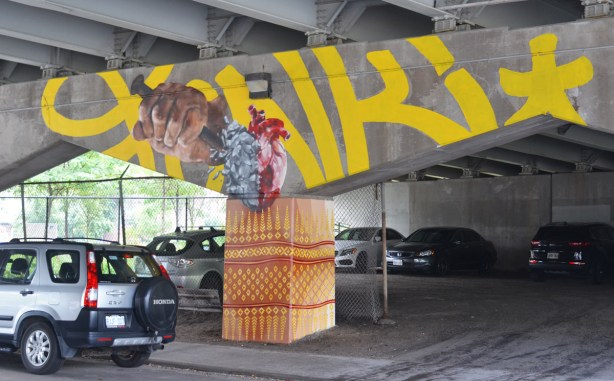 A concrete bent in an underpass that is a hand holding a large spike that is being plunged into a bleeding heart. Cars are parked underneath