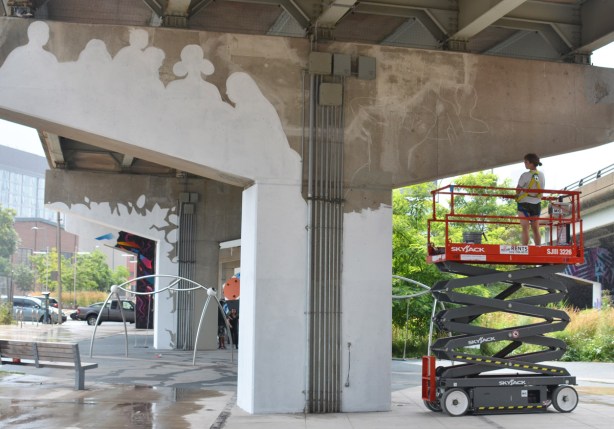 a person stands on an elevated platform and looks at the white outline that they have made in preparation for painting the bent white, the beginnings of a mural