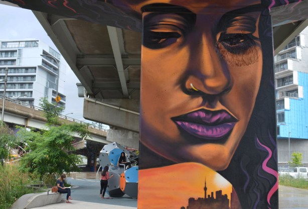 a mural of a woman's face in orange and purple tones, large, purple lips, on a concrete pillar, with young woman sitting in the park in the background