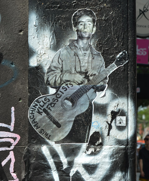 black and white paste up of Woody Guthrie standing, holding onto his guitar that has the words This machine kills fascists, written on it. 
