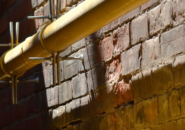 along an old brick exterior wall, there are two yellow gasline pipes that are held onto the wall with clamps
