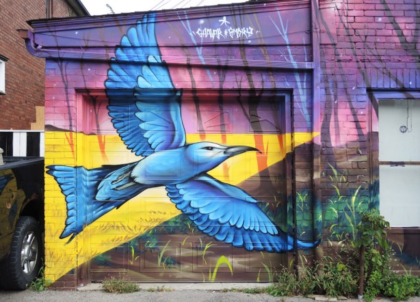 part of a larger mural by Clandestinos (Shalak Attack and Bruno Smoky) on the side of Riders bike store, a bluebird in flight painted on the garage door, yellow triangle of light behind it 