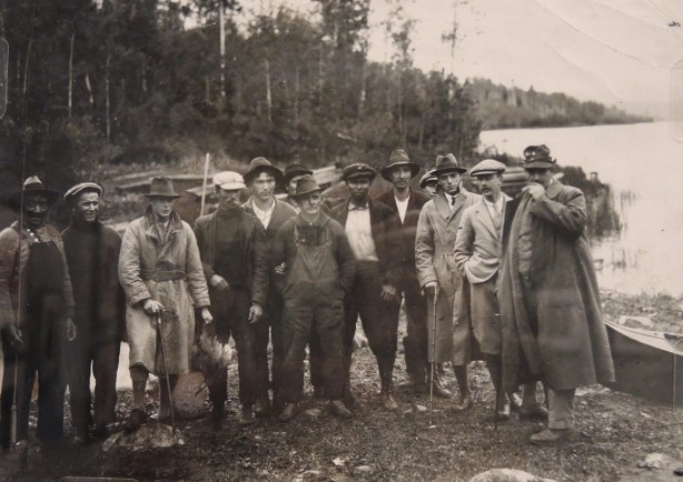 a vintage black and white photo of a group of men in northern Ontario, by a lake, one is holding a duck that has been shot