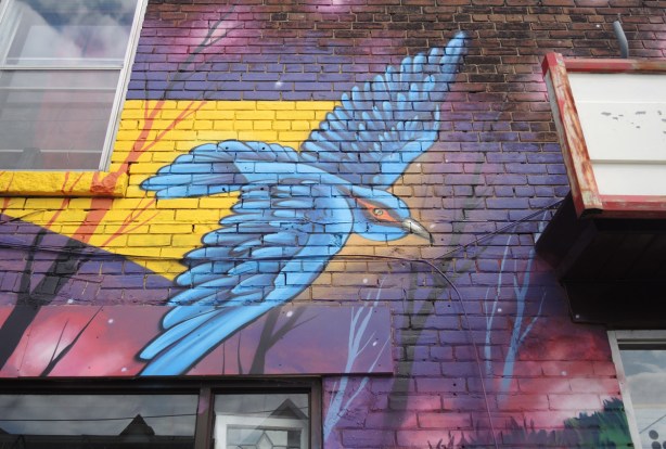 part of a larger mural by Clandestinos (Shalak Attack and Bruno Smoky) on the side of Riders bike store, a blue bird in flight with purplish coloured sky behind 