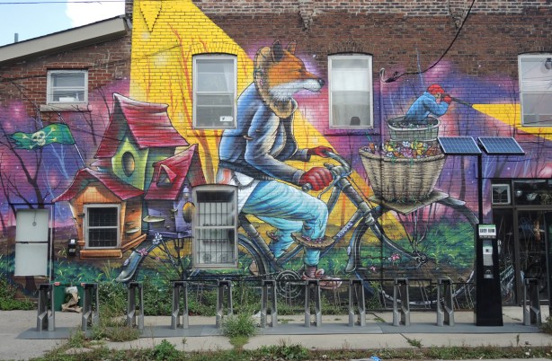 part of a larger mural by Clandestinos (Shalak Attack and Bruno Smoky) on the side of Riders bike store, main part of mural, a fox in jeans is riding a bike with a wicker basket in the front 