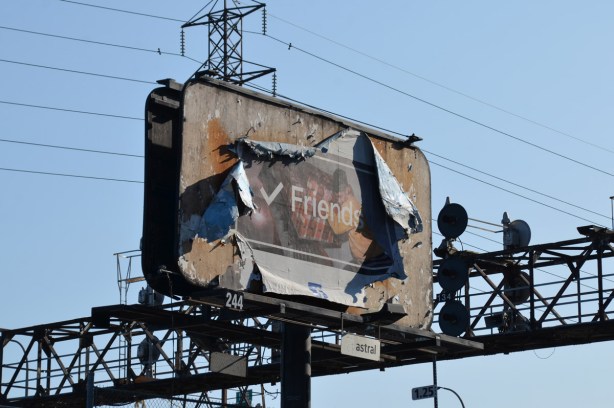 large billboard close to lights across the railway tracks, picture is badly peeling away from the edges. The picture in the middle is faded, but the word friend is written on top of it.