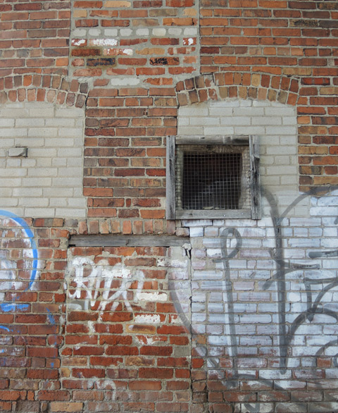 part of a brick wall that has old windows bricked over in a different brick, an old window with old wood frame, unpainted, some graffiti on the wall 