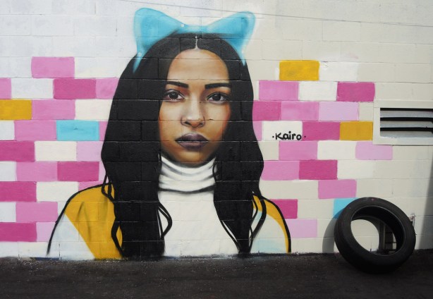 portrait of a woman on a wall, a mural by kairo, long dark hair, a blue bow on the top of her head, some blocks of the concrete block wall are painted in shades of pink 