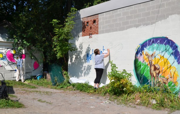 two women painting murals, Kim is in the background painting on a garage door while another woman is outlining a picture of a chameleon on a wall 