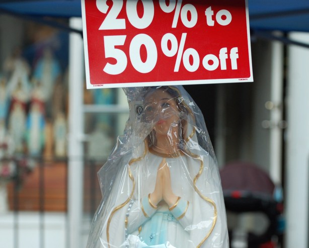 a statue of the VIrgin Mary wrapped in plastic, for sale at an outdoor sale, with a sign above her head that says 20% to 50 % off.