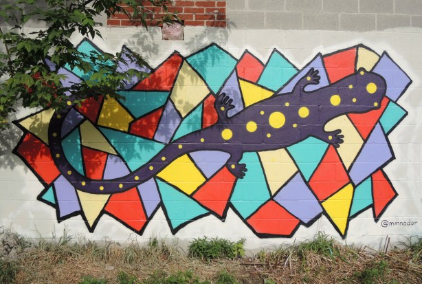 a dark purple salamander in a street art painting on a background of orange, yellow, and light blue polygonal shapes with 3 to 5 straight sides on a wall with a tree to the left
