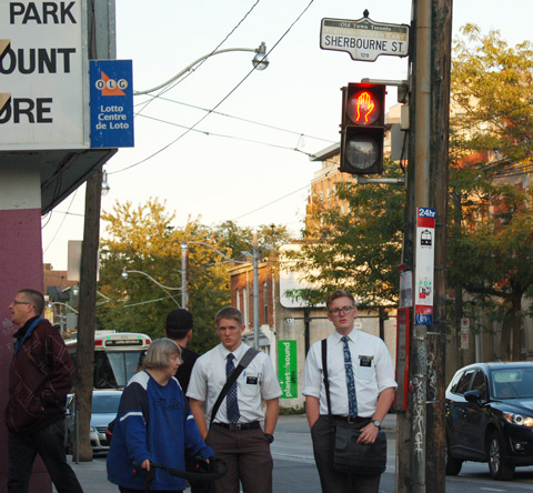 people standing on a corner waiting to cross the street, including a woman dressed in blue who is using a walker, plus two Morman men in their white shirts and black ties. 