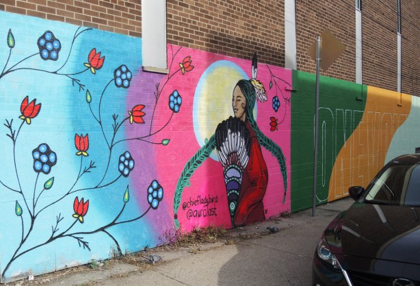 part of a mural by chief ladybird and aura of an indigenous woman with a feather in her hair, in front of the sun and circles of colour, red and blue flowers on the outer layer of colour (light blue) 