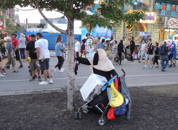 muslim woman in burka stands by a small tree with a stroller laiden down with parcels and bags, other people at the Ex are in the background