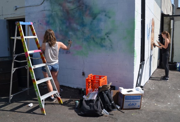 two women painting a mural, one mural each, one on the back of a building in an alley and the other on the side of the same building. 
