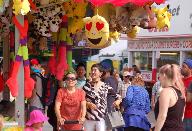 people walking past games atthe CNE, stuffies hanging from the roofs of game stalls 