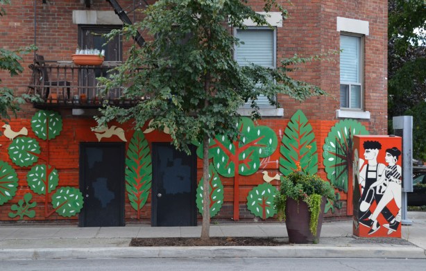 lower part of a brick building has been covered with a mural that is orange background, and cut out (wood) flat green trees in various shapes. A real tree grows in front of it, a metal traffic box is painted with a scene of two people walking with the same orange background