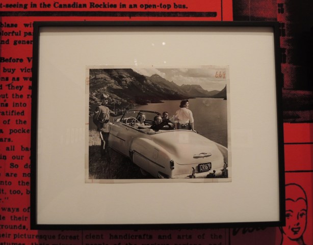 photo in an exhibit of a group of tourists in a convertible car with California plates parked beside the road and overlooking a mountain lake. An RCMP officer stands beside the car. 