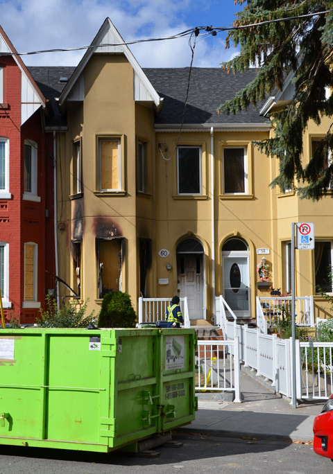 A row of houses where one is damaged from a recent fire.  Burned front door.  Windows have been boarded over, a skip for garbage isin front, workmen on the site 