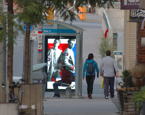 a lit advertisement in a bus shelter of two men in Roots clothes, a young man sits on the bench in the bus shelter while two people walk past it on the sidewalk