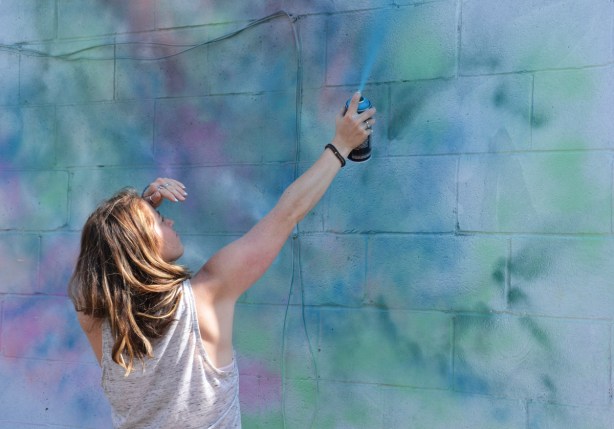 a woman sprays paint, light blue, onto the background of a mural that she's painting, blue, green and pinks that merge together with no defined lines or edges 