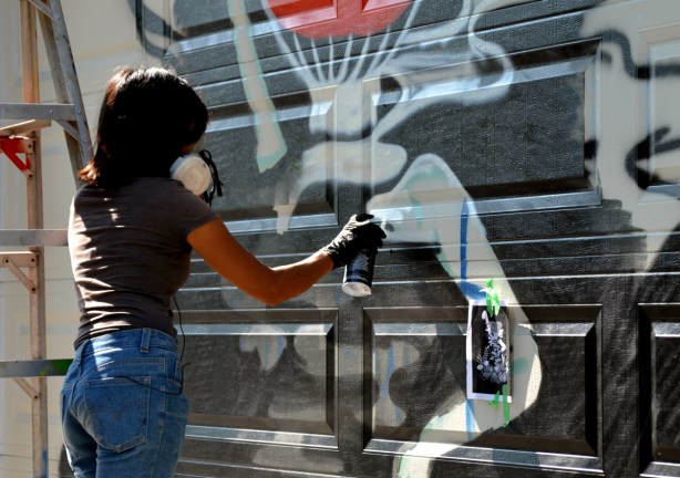 woman spraing black on a black and white mural, standing beside a ladder, on a garage door in an alleyy paint