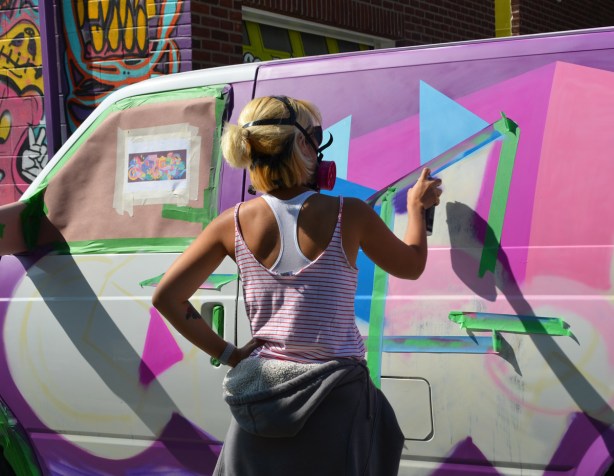 a woman spray paints on the side of a van that is being covered with street art, masking tape to keep the spray paint in straight lines. 