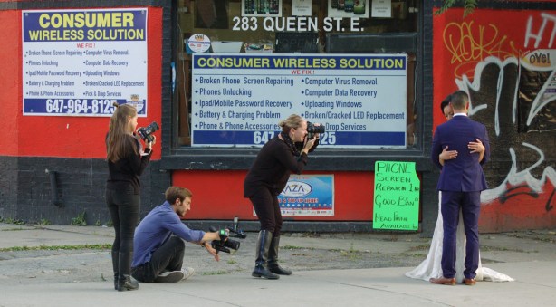 a bride and groom embrace while three photographers take their picture. on a sidewalk of a city street
