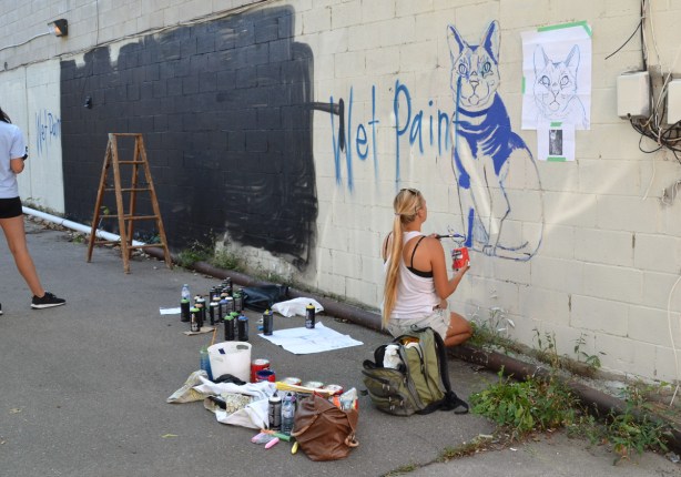 a woman is crouching beside a wall where she is painting a picture of a blue cat, on the wall beside her is a large black section that has just been painted in preparation for another mural