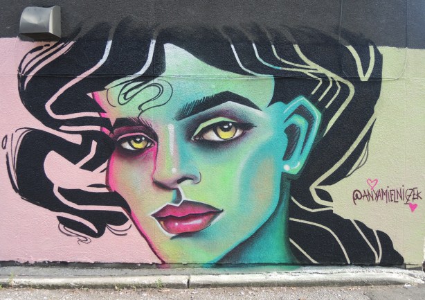 street art mural of a woman's face in greens and blues on one side, moving to pink on the other, lots of black hair. red lips, yellow eyes