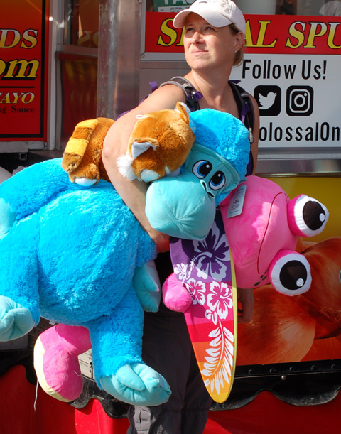 a woman carrying two stuffies and a skateboard, one is a large blue dinosaur and the other is a large pink creature