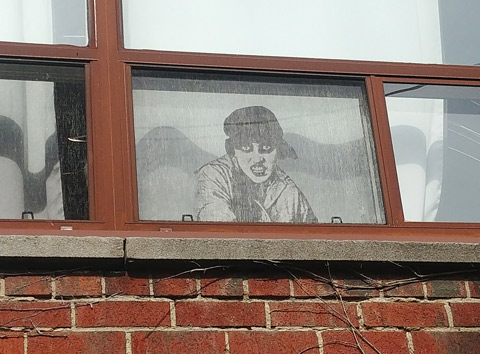 black and white realistic drawing of a sneering woman with a hat on looking out of a window on the second storey of a brick building. 