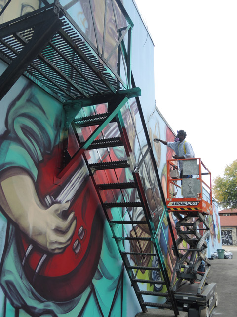 elicser works on painting a mural, in the foreground is a large painted guitar player with a red uitar, painted behind a metal fire escape staircase 