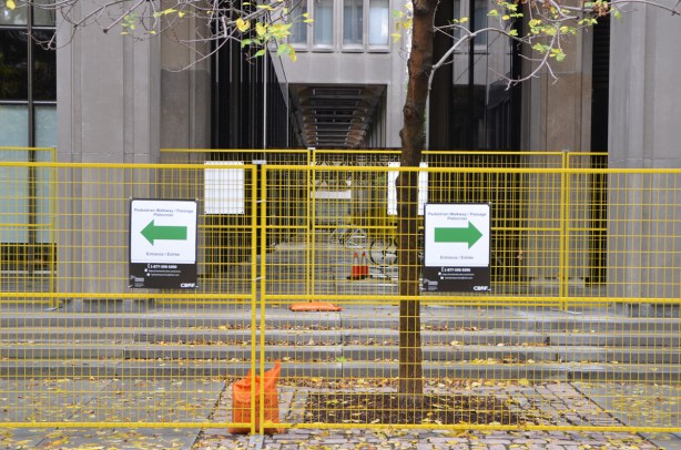 a yellow construction fence in front of a stairs between two buildings, two signs on the fence, both are green arrows but they point in opposite directions. 