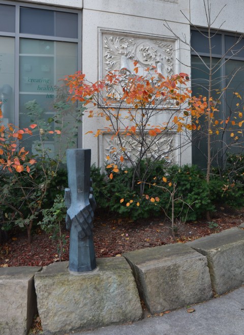 a small sculpture of two hands holding, vertical, one hand from above and the other from below, in a small garden with some shrubs with autumn foilage, orange colours