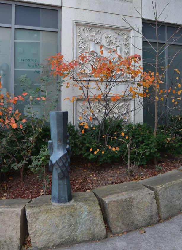 a small sculpture of two hands holding, vertical, one hand from above and the other from below, in a small garden with some shrubs with autumn foilage, orange colours 