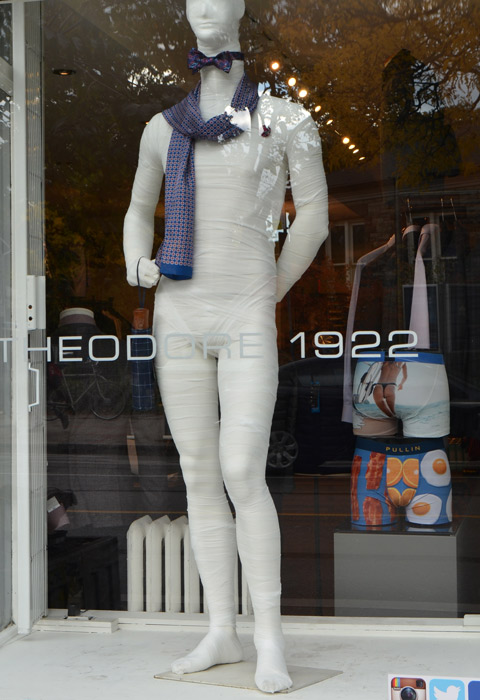 a mannequin in a window of a men's clothing store, Theodore 1922, wrapped up like a mummy, scarf loosely draped around his neck, wearing a bowtie, and holding a folded up compact umbrella