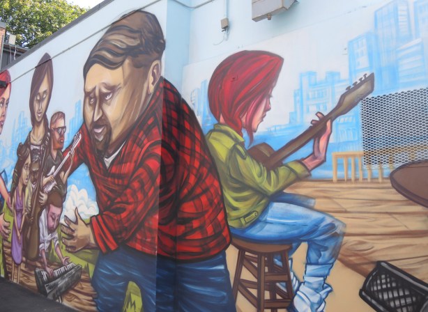 part of a mural by elicser of people - a woman is sitting on a stool and playing a guitar, a man in a red and black checked shirt 